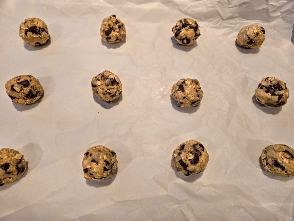 Twelve unbaked oatmeal chocolate chip cookie dough balls are arranged on a sheet of parchment paper in a grid pattern, ready to be baked.