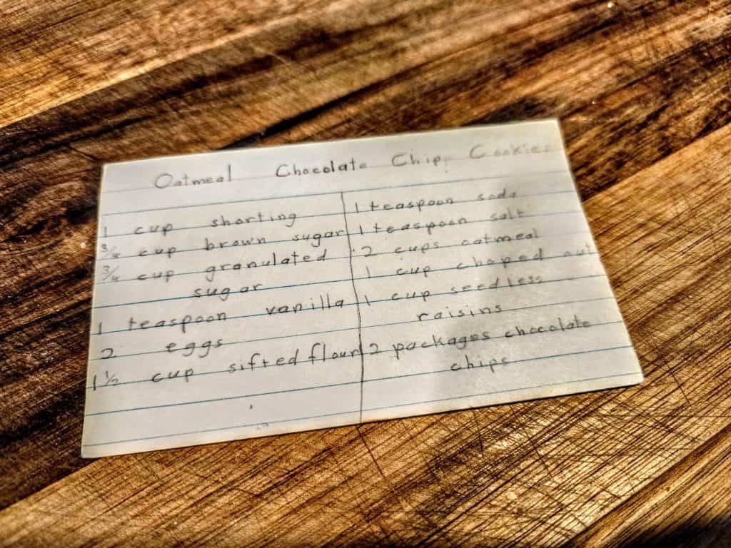A handwritten recipe card titled "Grandma's Oatmeal Chocolate Chip Cookies" lists ingredients like shortening, sugars, eggs, vanilla, flour, oatmeal, soda, salt, raisins, and chocolate chips. This cherished card rests on a wooden surface.