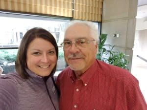 A woman with shoulder-length brown hair wearing a purple top stands next to a man with white hair and glasses, wearing a red shirt. They are smiling indoors, framed by large windows and plants, sharing a moment of love that hints at the legacy of their shared journey.