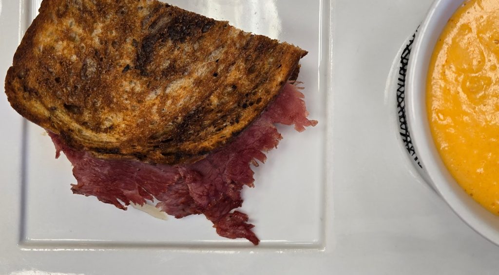 A Reuben sandwich with corned beef on toasted bread sits elegantly on a square white plate, beside a bowl of creamy orange soup, offering a taste reminiscent of the delightful dining experiences in Hays, KS.