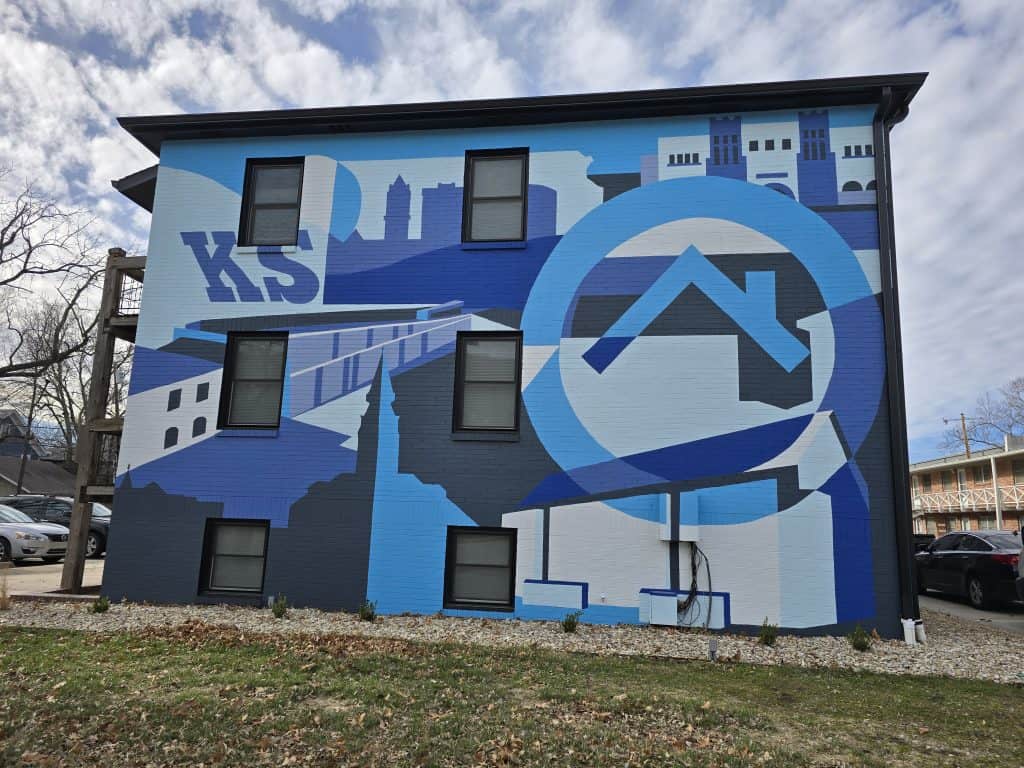 A building wall features a mural with geometric shapes and designs in blue tones. Elements include abstract buildings, the letters "K S," and a stylized house. The sky is partly cloudy, and the ground has some dry leaves.