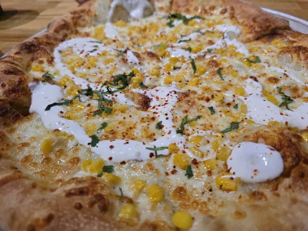 Close-up of a pizza topped with melted cheese, corn, white sauce, red pepper flakes, and chopped herbs on a wooden surface at Defiance Brewing Company in Hays KS.