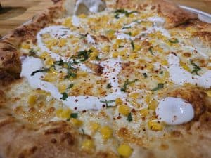 Close-up of a pizza topped with melted cheese, corn, white sauce, red pepper flakes, and chopped herbs on a wooden surface at Defiance Brewing Company in Hays KS.