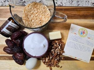 A wooden cutting board with ingredients for a Date Ball Recipe: a bowl of Rice Krispies, vanilla bean paste, whole dates, sugar, pecans, an egg, butter, and a handwritten recipe card&mdash;just like Grandma's Recipe.
