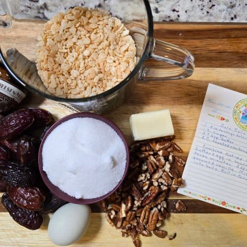 A wooden cutting board with ingredients for a Date Ball Recipe: a bowl of Rice Krispies, vanilla bean paste, whole dates, sugar, pecans, an egg, butter, and a handwritten recipe card—just like Grandma's Recipe.