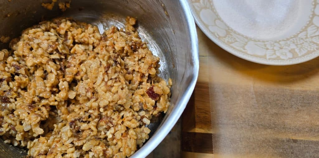 A metal bowl filled with a sticky mixture of puffed rice and nuts—just like Grandma's Date Ball Recipe—sits on a wooden surface next to a white patterned plate.