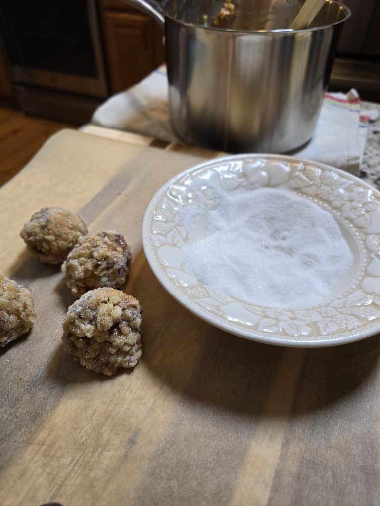 Three unbaked cookie dough balls rest on a wooden surface next to a decorative white plate filled with granulated sugar, ready to be rolled into classic date balls inspired by grandma's recipe, with a pot and wooden spoon in the background.