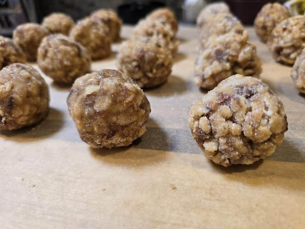 Close-up of several round oatmeal cookie dough balls, inspired by Grandma's Date Ball Recipe, with visible oats and sugar on a parchment-lined surface. More dough balls are slightly out of focus in the background, ready for baking.