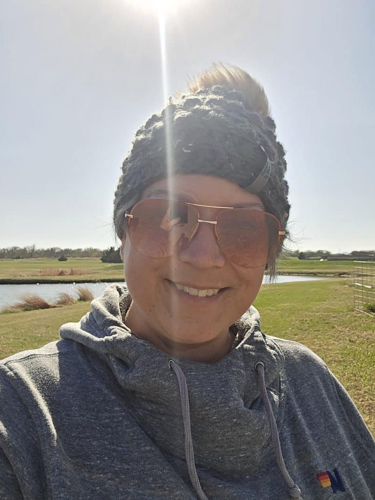 A person wearing sunglasses, a grey headband, and a grey hoodie smiles outdoors under bright sunlight, enjoying life after chemotherapy with a green field, a pond, and clear sky in the background.