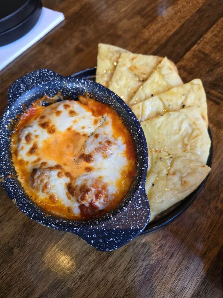 At Bootsie's in Manhattan KS, a black speckled bowl filled with cheesy baked meatballs in tomato sauce is served alongside slices of toasted flatbread on a wooden table.