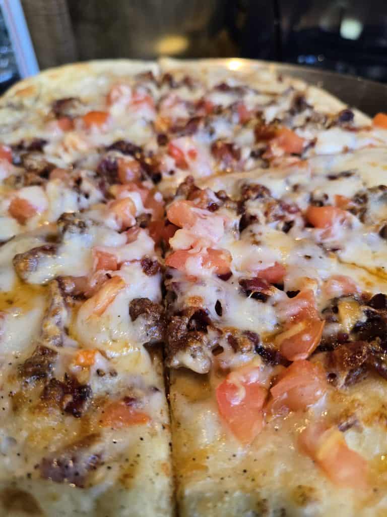 Close-up of a freshly baked Bootsie's pizza in Manhattan KS, topped with melted cheese, ground meat, bacon bits, and diced tomatoes, with a sliced piece in the foreground.