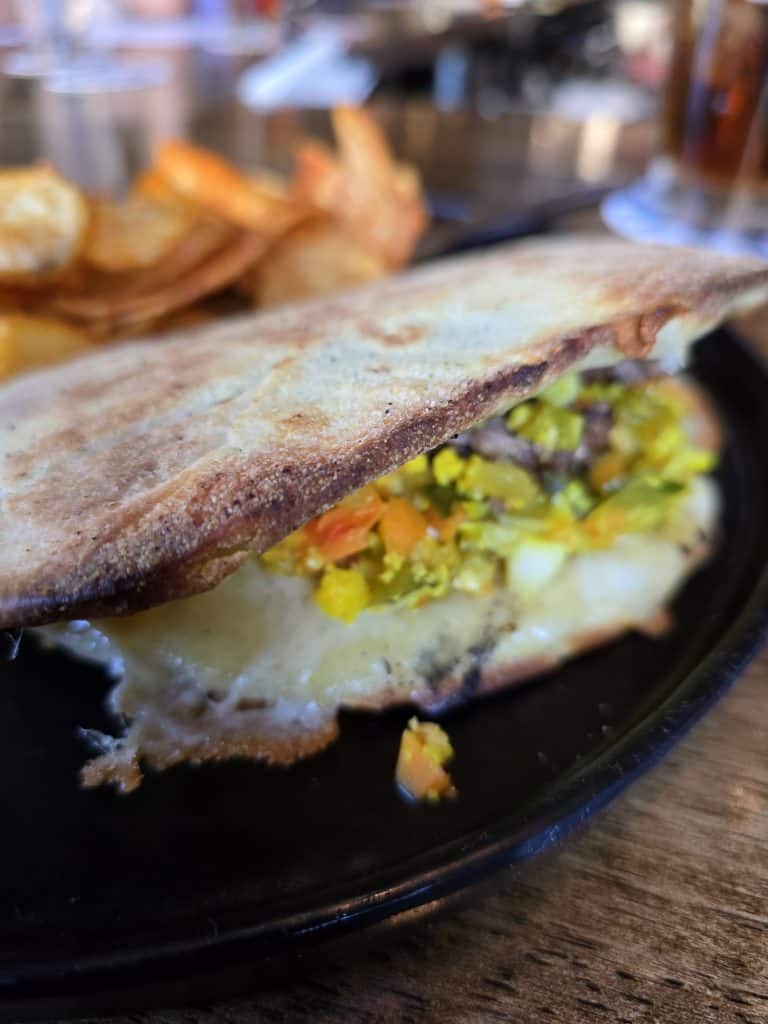 A close-up of a sandwich with melted cheese and chopped vegetables on a black plate, with potato chips in the background on a wooden table—a tempting bite inspired by favorites from Manhattan restaurants like Bootsie’s in Manhattan, KS.