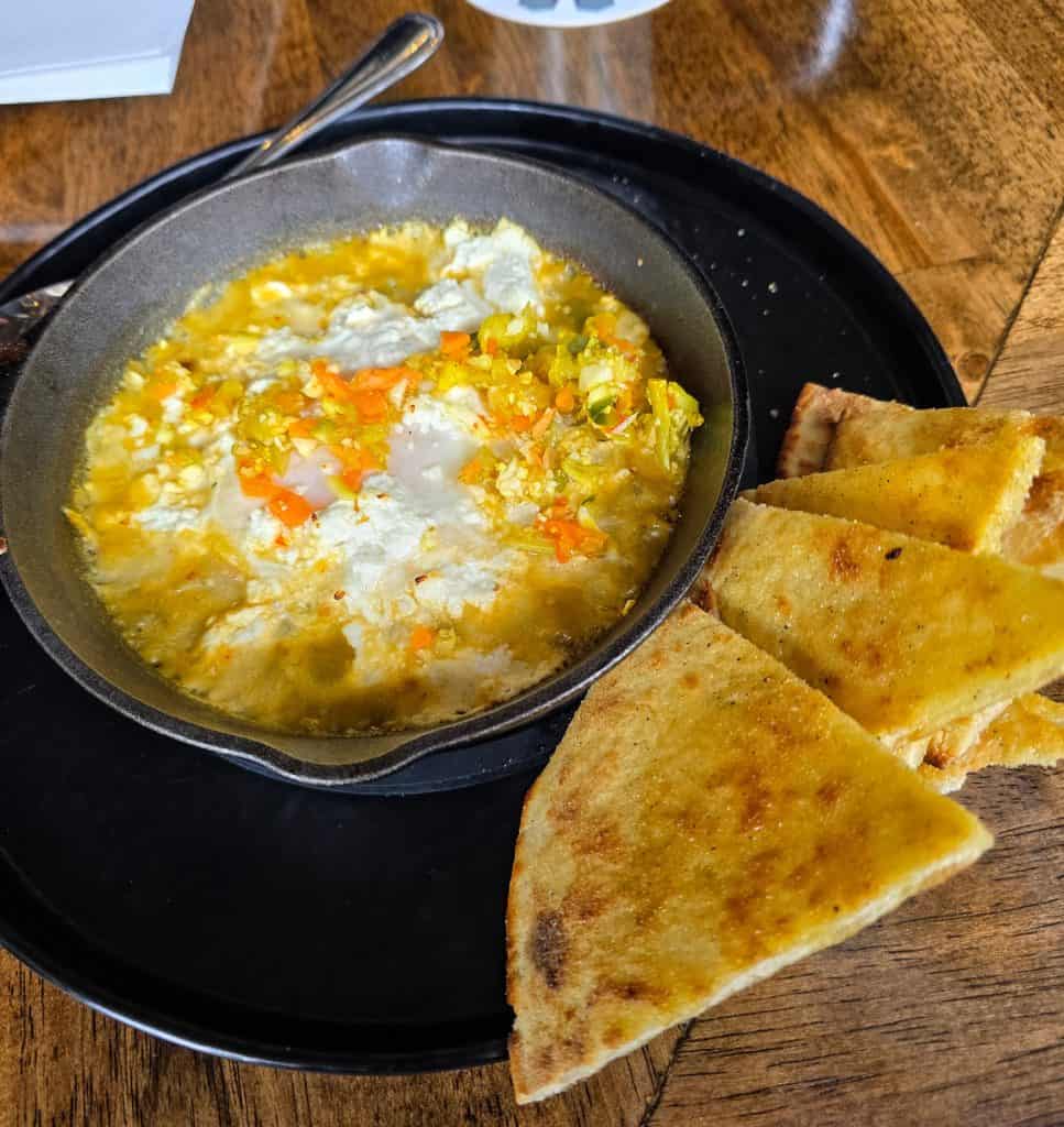 A black plate holds a skillet of eggs cooked with vegetables next to several triangular slices of golden, toasted flatbread at Bootsie's in Manhattan KS, all served atop a rustic wooden table.