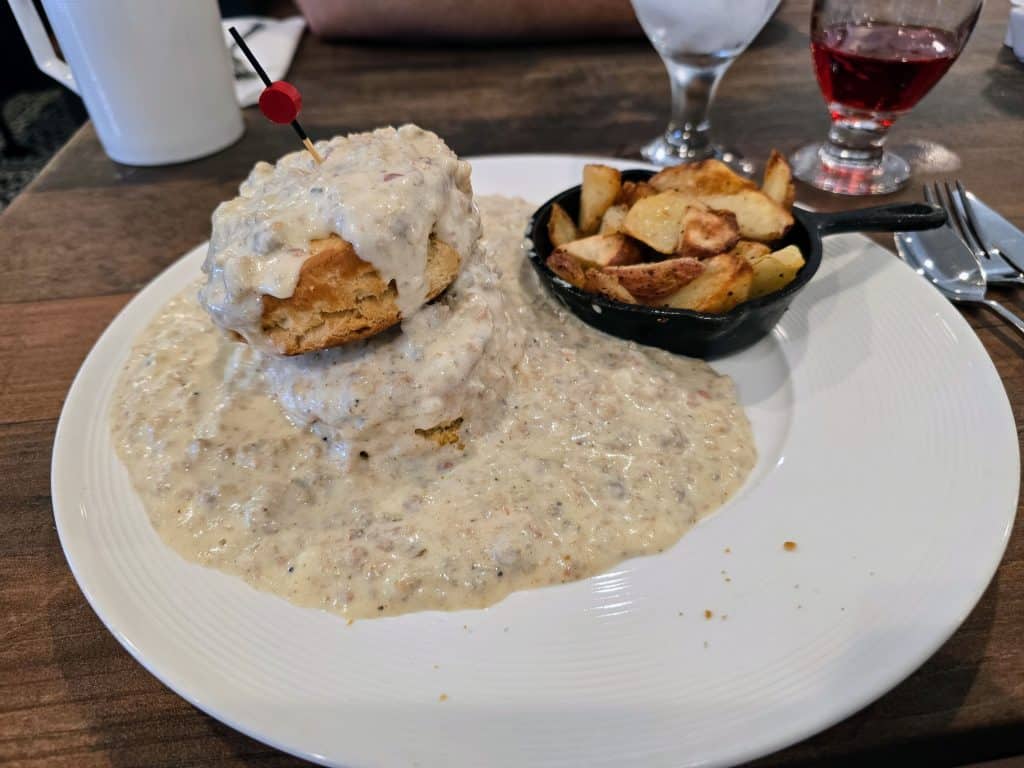 At Breaks Eatery & Bar in Hays KS, a plate of biscuits smothered with creamy sausage gravy pairs perfectly with a small skillet of roasted potato wedges. A water glass and a red drink sit nearby on the wooden restaurant table.