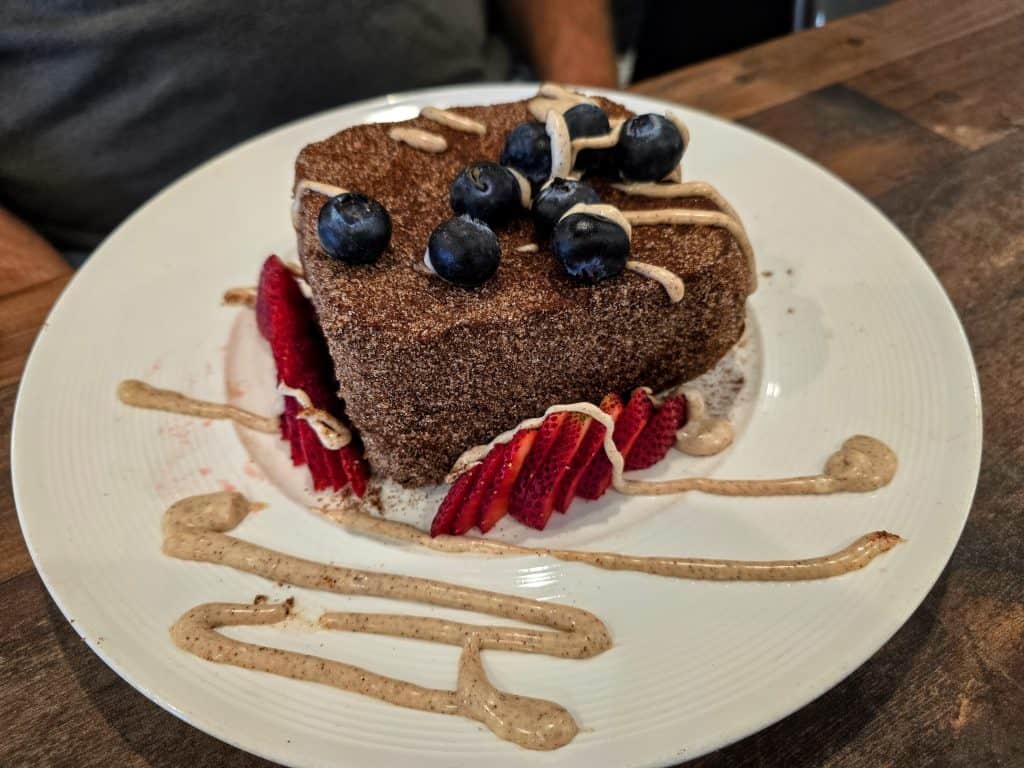 A heart-shaped chocolate cake topped with blueberries and surrounded by sliced strawberries sits on a white plate at Breaks Eatery & Bar, Hays KS. Drizzled with a light brown sauce, the dessert rests on a wooden table.