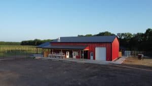 A bright red barn at Fall Color Farms with a covered porch, white chairs, and a large garage door sits beside a field and trees under a clear blue Riley KS sky.