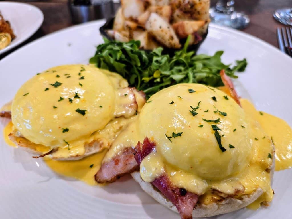 Two eggs Benedict topped with creamy hollandaise sauce and sprinkled with herbs, served on toasted English muffins with bacon. Enjoy seasoned potatoes and fresh greens at Breaks Eatery, a must-visit Hays restaurant.