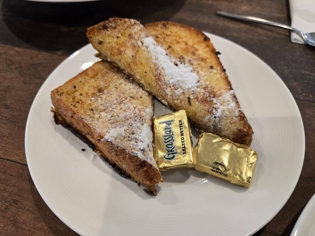 Two slices of toasted bread with powdered sugar on top, served on a white plate with two gold-wrapped packs of salted butter, at Breaks Eatery & Bar in Hays KS, set on a wooden table.