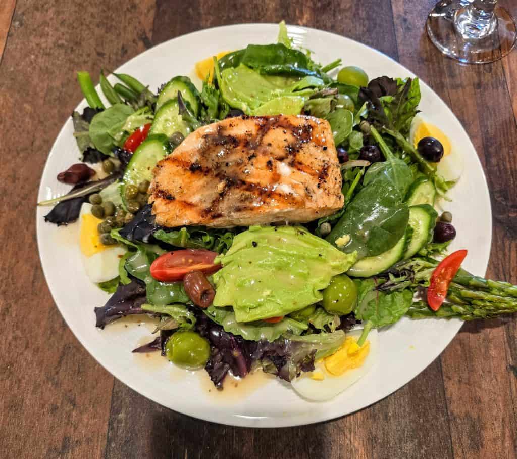 A plate of mixed green salad topped with grilled salmon, avocado, cherry tomatoes, cucumber, olives, capers, asparagus, and hard-boiled eggs awaits you at Breaks Eatery & Bar in Hays KS—your go-to restaurant for fresh flavors.