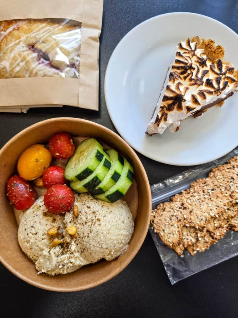 A bowl with hummus, cherry tomatoes, and cucumber slices sits next to a bag of pastry at Hungry Chickpea restaurant in Manhattan KS, alongside a plate with meringue pie and several pieces of seeded crackers on a dark surface.