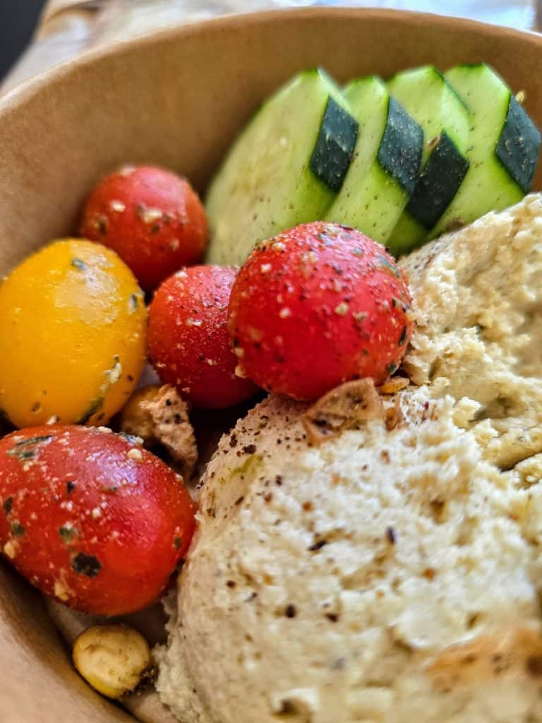 A close-up of a bowl at Hungry Chickpea restaurant in Manhattan KS, featuring sliced cucumber, red and yellow cherry tomatoes, and a scoop of herbed hummus, all garnished with seasoning and roasted garlic.