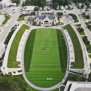 Aerial view of a stadium with green turf, marked for multiple sports, at Kansas State University. Surrounded by stands with grassy rooftops, a few people enjoy the field as buildings and roads encircle the campus landmark.