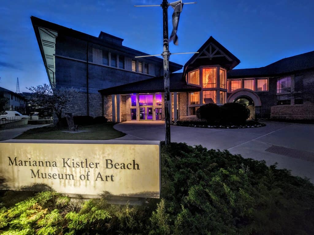The Marianna Kistler Beach Museum of Art building at dusk, warmly lit from within, stands on the K-State Campus in Manhattan KS, with a sign in the foreground and landscaped greenery surrounding the entrance.