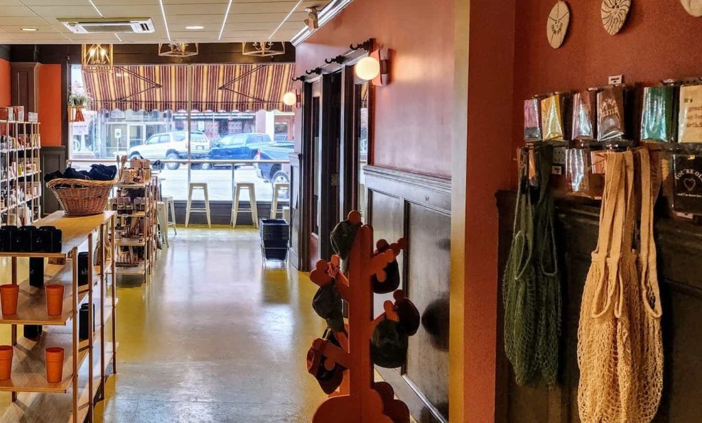 A view inside a small shop in Manhattan KS with refrigerated displays on the left, shelves of goods in the center, and large windows at the front letting in daylight—one of the most Instagrammable spots near the scenic Flint Hills. Tables and chairs sit by the entrance.