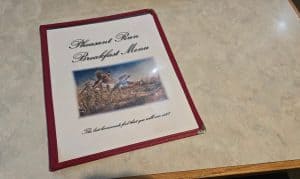A laminated Pheasant Run breakfast menu rests on a light table in Hays, KS. Its red border frames an image of pheasants in a field, while a slogan highlights their homemade food.