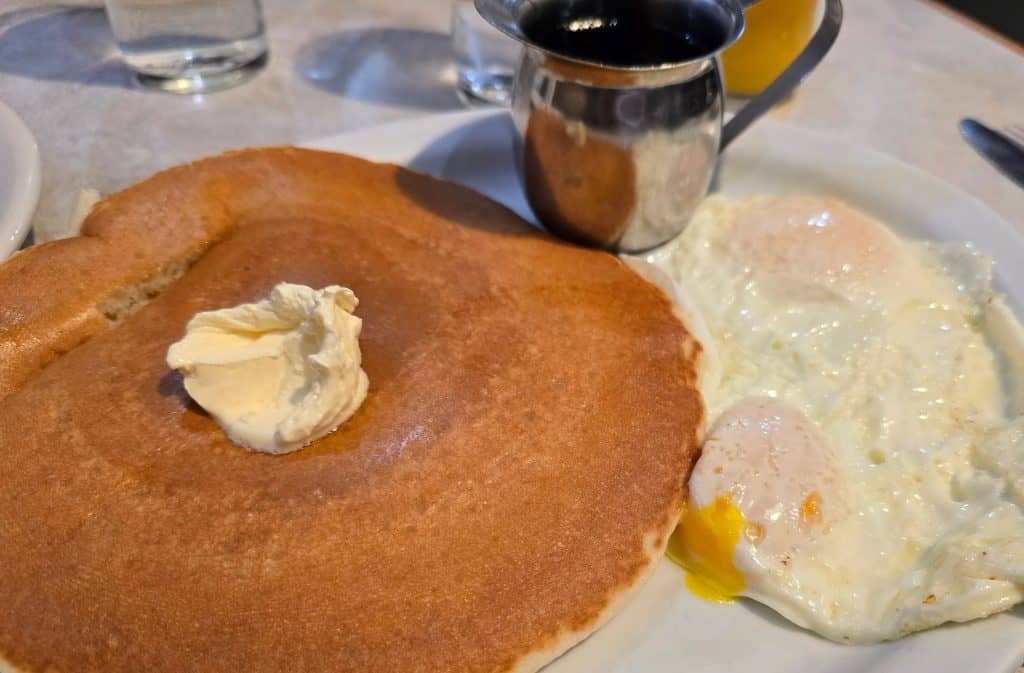 A breakfast plate at Pheasant Run in Hays, KS features a large pancake with butter, two sunny-side-up eggs, and a metal cup of syrup. A glass of water and another drink can be seen in the background.
