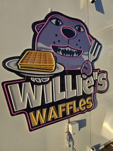 A cartoon animal holding a plate with waffles and a fork is featured above the text "Willie's Waffles" in bold, colorful letters on a white background, capturing the fun spirit of Willie's Waffles in Manhattan KS.