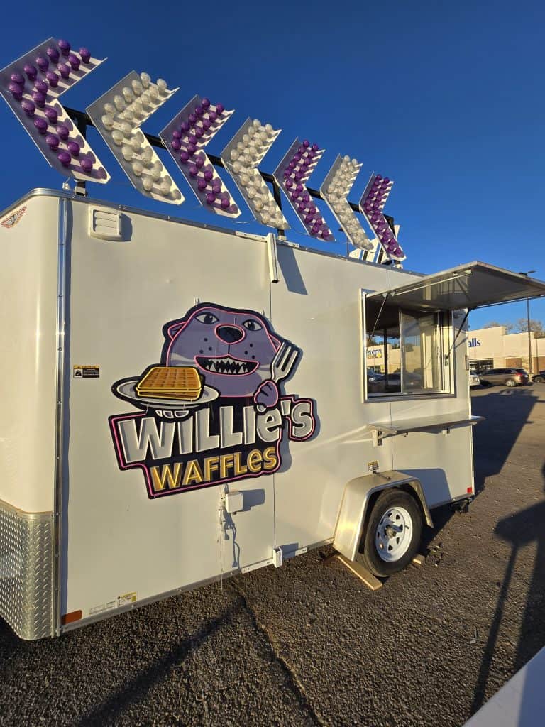 A white food trailer in Manhattan KS features a cartoon dog holding waffles and the text "Willie's Waffles" on the side. Colorful arrow-shaped lights line the roof as it sits parked in a sunny lot.