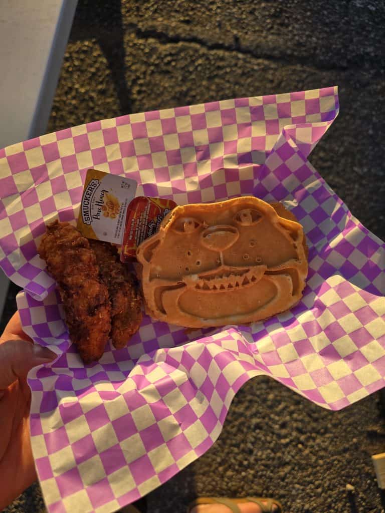 A hand holds a checkered tray from Willie's Waffles in Manhattan KS, featuring a bear-shaped waffle, two fried chicken strips, Smucker's honey, and grape jelly—all served on purple and white checkered paper.