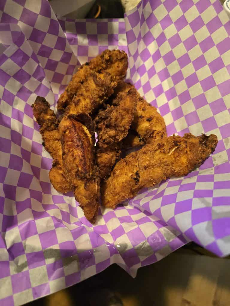 A serving of crispy fried chicken tenders is placed on a sheet of purple and white checkered paper in a basket, perfect to pair with hot waffles from Willie's Waffles in Manhattan KS.