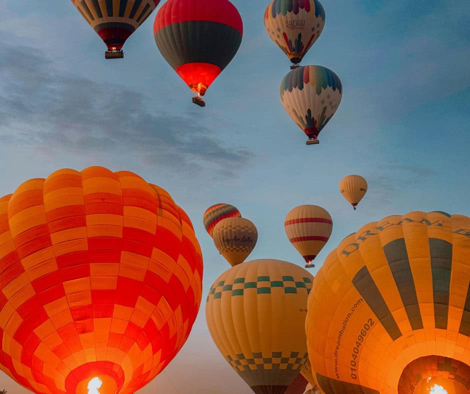 A group of colorful hot air balloons rises into the sky at sunrise during the Flint Hills Balloon Festival in Manhattan KS, with several close to the camera and others floating higher against a blue and orange-tinged sky.