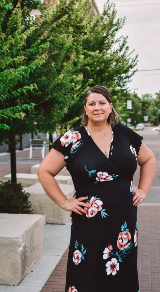 A woman with brown hair stands confidently on a sidewalk in Manhattan KS, wearing a black dress with a floral pattern. She has her hands on her hips, posing at one of the city's most Instagrammable spots, with trees and buildings in the background.