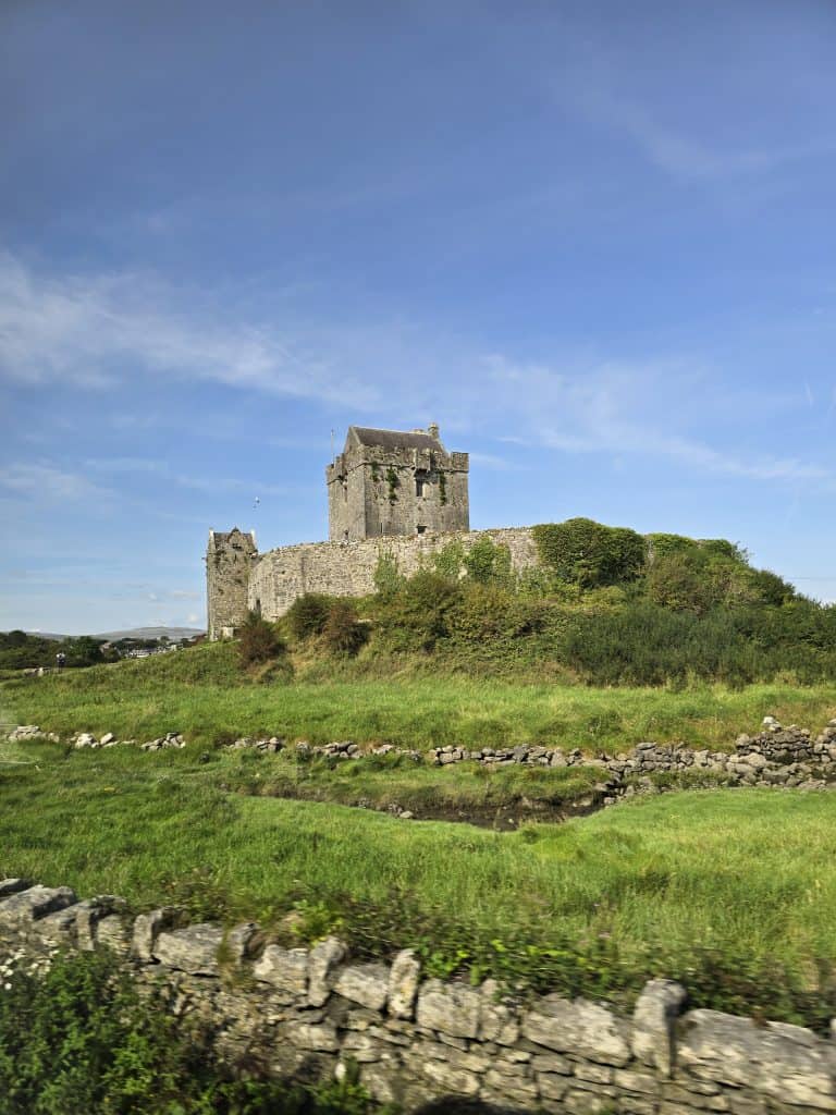 A stone castle with a square tower stands atop a grassy hill, surrounded by stone walls and lush greenery—a perfect sight on your Ireland trip under a clear blue sky.