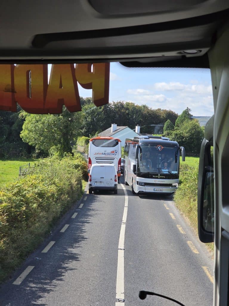 During a trip to Ireland, a narrow country road becomes a tight squeeze as two buses and a white van try to pass each other. The photo, taken from inside another vehicle, captures trees and grass lining this classic Ireland travel scene.