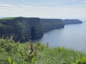 The Cliffs of Moher in Ireland rise steeply above the Atlantic Ocean, with green grass and wildflowers in the foreground&mdash;a must-see on any Ireland vacation under a partly cloudy sky.