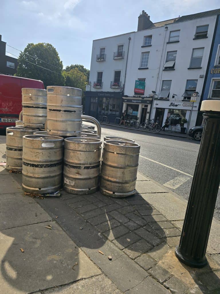 Several metal beer kegs are stacked on a sidewalk near a street lined with white buildings, storefronts, and parked bicycles on a sunny day—perfect for capturing the charm of an Ireland vacation or your next Ireland trip. A red van is partially visible on the left.