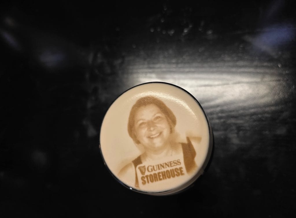 A pint of Guinness with a foam top printed with a smiling woman's portrait and the words "Guinness Storehouse"—a must-see for any Ireland trip—stands out against a dark background.
