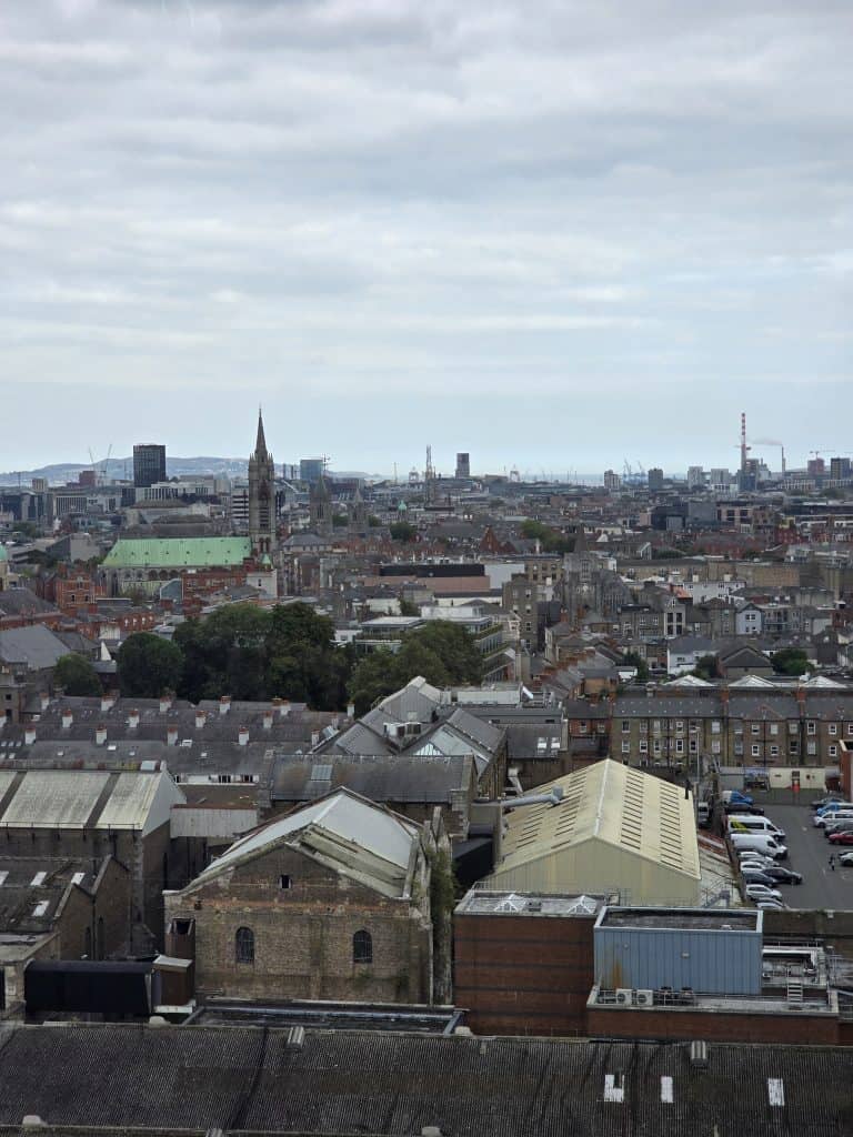 A cityscape with rows of old brick buildings, a church spire, patches of greenery, and an overcast sky—perfect for those looking to travel Ireland. Industrial cranes and distant hills complete this authentic Irish scene.