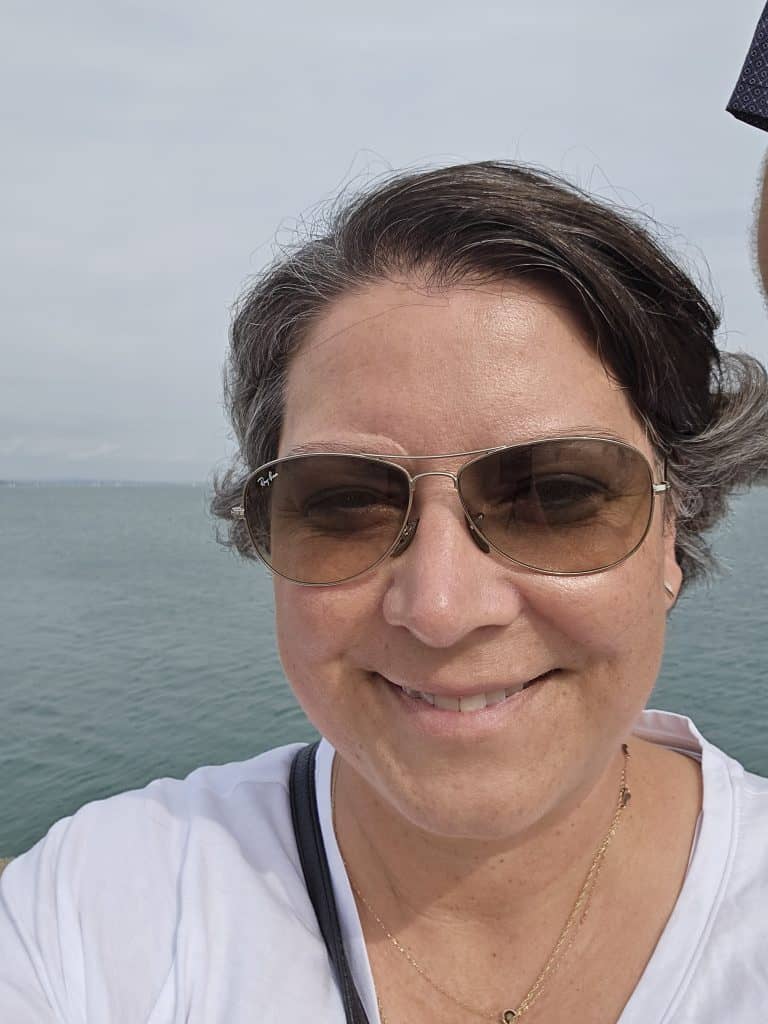 A woman wearing sunglasses and a white shirt smiles at the camera, with a calm, blue body of water and cloudy sky in the background—perfect inspiration for your next Ireland travel adventure.