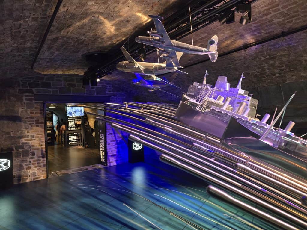 A highlight for Ireland tourism, this museum exhibit features a large model warship and an airplane above blue waves, set beneath a brick arched ceiling and leading to another room with illuminated displays—a must-see on your Guide to Ireland.
