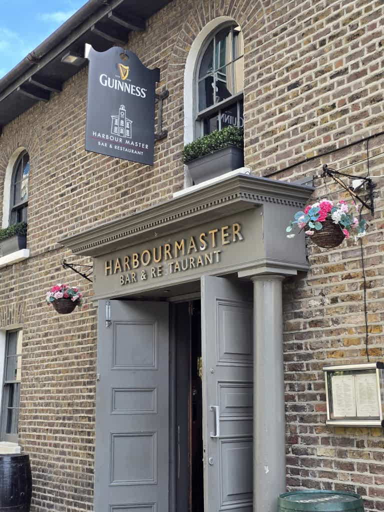 Entrance of the Harbourmaster Bar & Restaurant, a brick building with grey doors, hanging flower baskets, and a Guinness sign above the door—a charming spot often featured in Ireland travel guides. A menu is displayed on the wall next to the entrance.