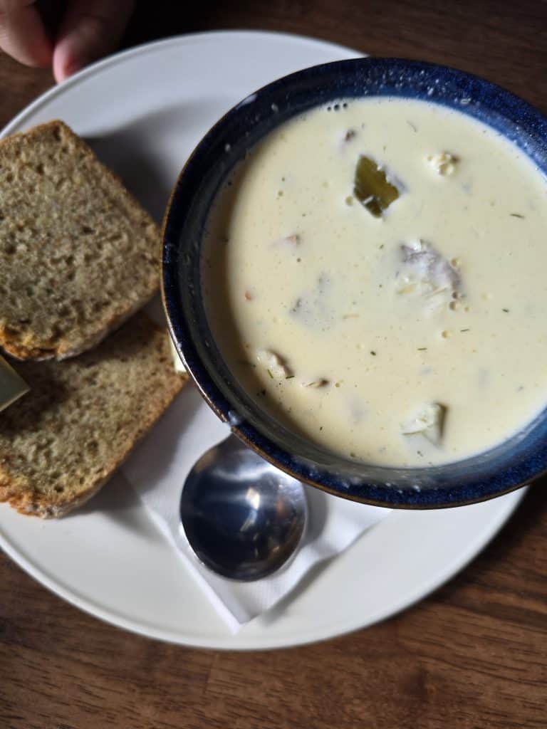 A bowl of creamy soup with visible herbs and chunks, served on a white plate with two slices of brown bread and a small pat of butter—a classic taste of Ireland, perfect for any travel guide or tourism experience, alongside a metal spoon.