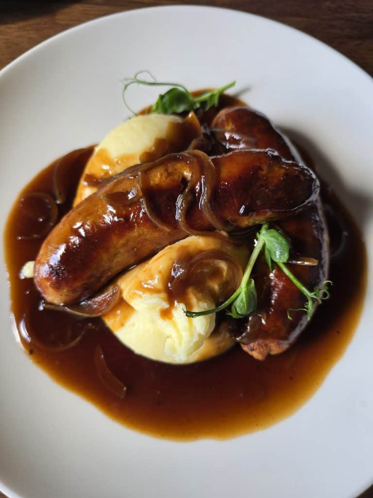 A classic taste of Ireland: two sausages on creamy mashed potatoes, topped with rich brown onion gravy and fresh pea shoots—perfect for any travel guide or tourism feature on authentic Irish cuisine.