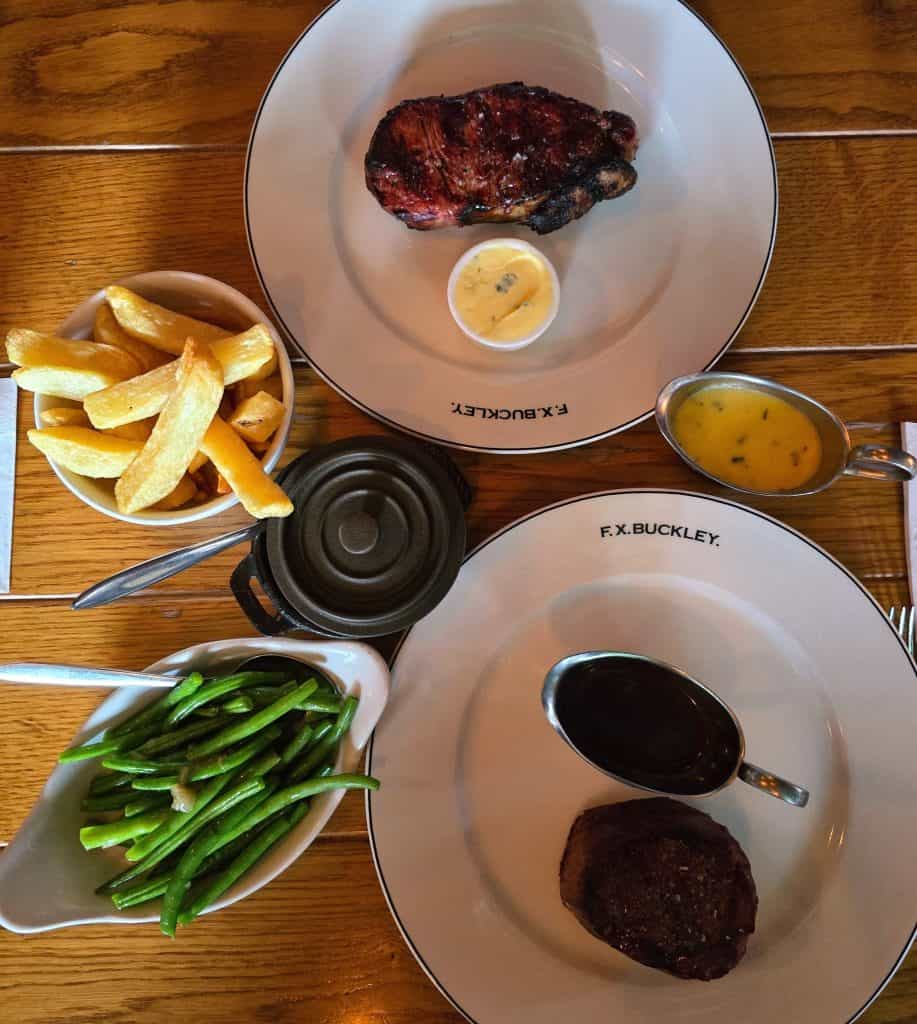 Two plated steaks with sauces are served on a wooden table, accompanied by thick-cut fries and green beans. The "F.X. Buckley" plates evoke classic Irish dining—a must-try spot for anyone looking to travel Ireland and explore its cuisine.