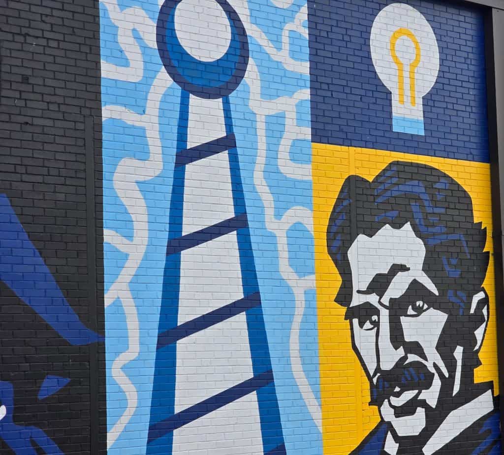 A colorful building mural in Hays, KS features a stylized portrait of Nikola Tesla, a large radio tower with radiating waves, and a glowing light bulb on a blue and yellow background for STEM Harvest.