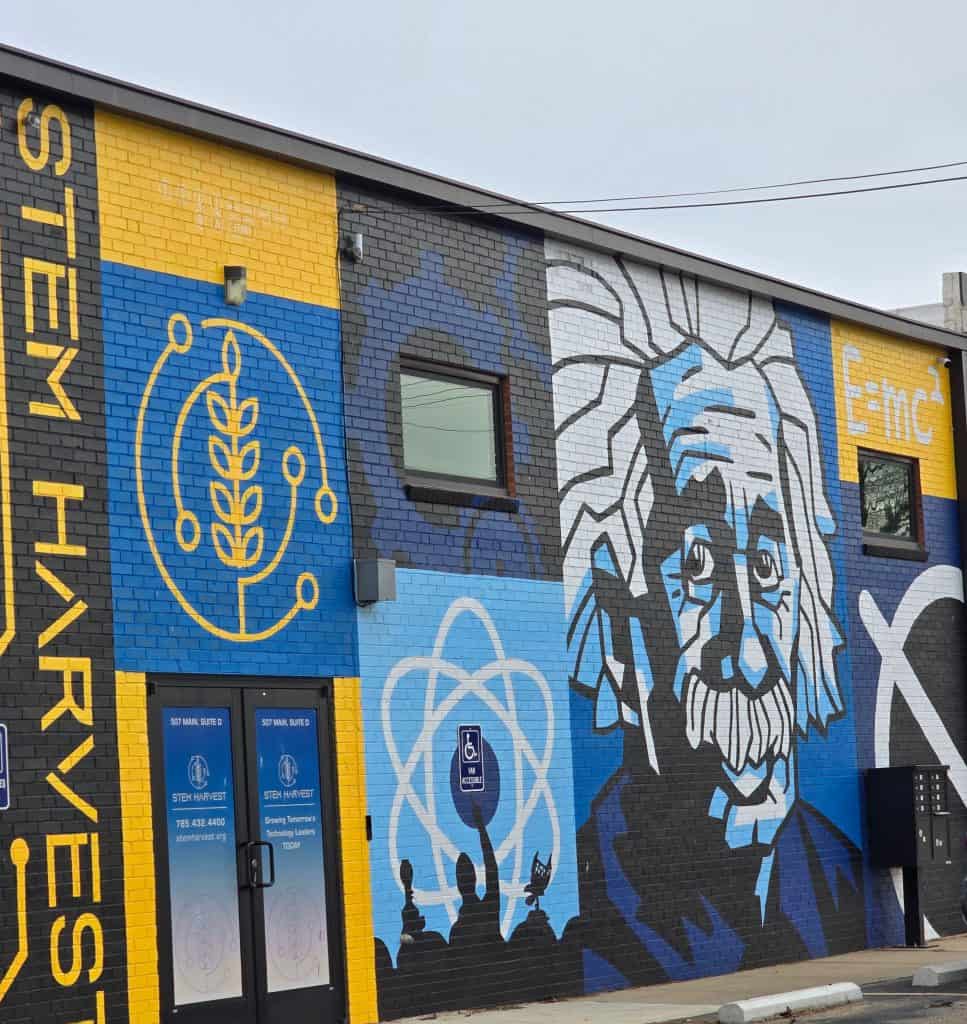 A colorful building mural in Hays, KS features scientific symbols, the equation E=mc², and a large portrait of Albert Einstein. The words "STEM HARVEST" are painted vertically on the left side of the brick building.